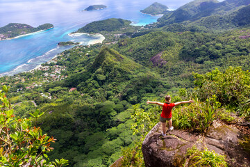 Young female traveller standing at the edge of the cliff at Morne Blanc View Point, overlooking Mahe Island coastline with lush tropical vegetation and crystal blue ocean, Seychelles. - obrazy, fototapety, plakaty