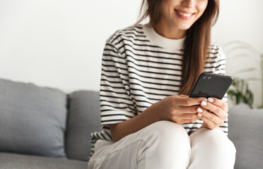 Cropped shot of girl smiles, sits at home and uses smartphone. Young woman chatting on mobile phone, sitting on sofa living room, using cellphone