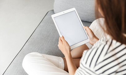 Cropped shot of woman, hands holding digital tablet with white screen, mockup promotion empty space on display, sitting with crossed legs on sofa at home