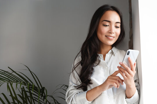 Image of young asian business woman standing near window with smartphone. Girl chat in application, answer phone call while working from home, sending smth via mobile phone app