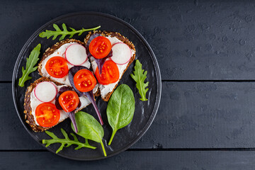 Toasts with cream cheese, cherry tomatoes and radish on black plate. Sandwiches with cream cheese and fresh herbs on a dark boards. Copy space. Flat lay