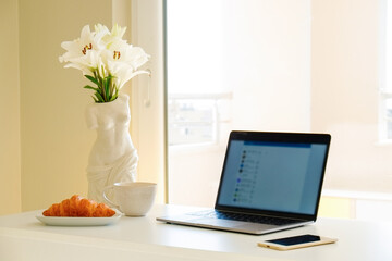 Home office concept. Designated work from home area near the window. Modern laptop, notebook and vase with flowers on table. Close up, copy space, background.