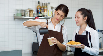 Young caucasian barista pouring milk from the jug into a paper coffee cup. Asian assistant in an apron holding croissant plate stands beside her. Morning atmosphere in a coffee shop.