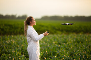 female agricultural specialist holding Drone Remote and controlling drone in air standing in corn...