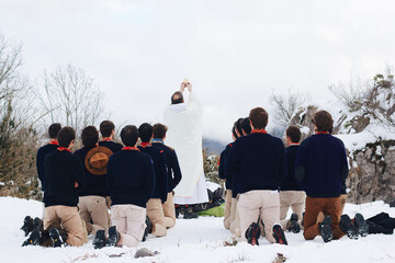 Rear view of a group of men kneeling on the snow with a priest in white standing in front
