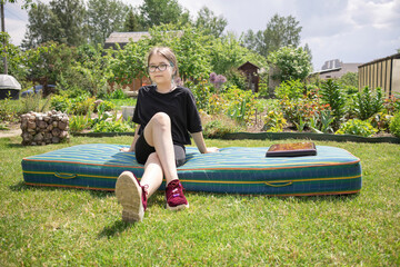 A teenage girl in glasses with multi-colored hair sits on a mattress in a blooming summer garden, next to a closed laptop