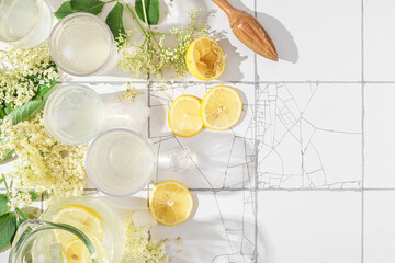 Fresh iced mocktail or lemonade with lemons and elderflowers in glasses on white tile table with copy space. Top view