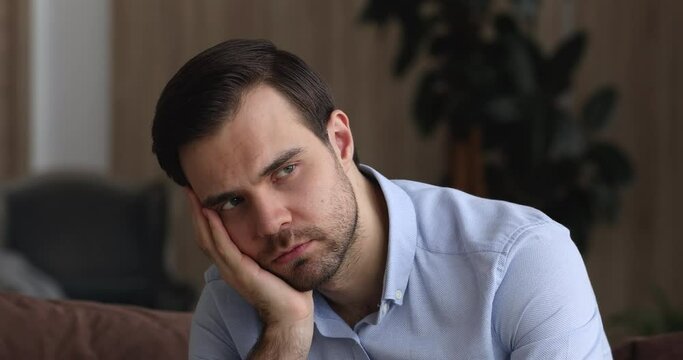 Sad bored man lean head on hand thinking seated on sofa at home alone, lost deep in thoughts feeling boredom, close up. Depressed troubled young guy reflecting feels lonely, melancholic mood concept