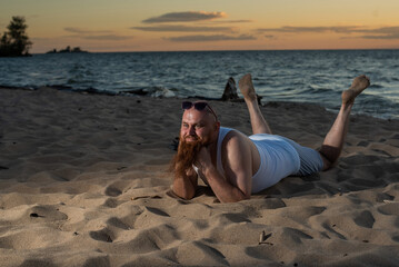 Funny bald man with red beard posing on the beach at sunset. A humorous male parody of a glamorous girl.