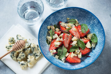 Salad with fresh strawberry, mangold leaves and blue cheese served in a blue bowl, high angle view on a light-blue stone background