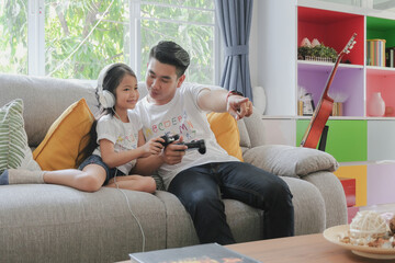 Obraz premium Two asian young father and adorable little girl daughter family enjoy playing game console together with happy smiling face at sofa living room on holiday weekend. Selective focus.