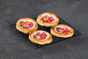 Small open pies made of puff pastry with ricotta and fresh strawberries on a serving rack on a dark...