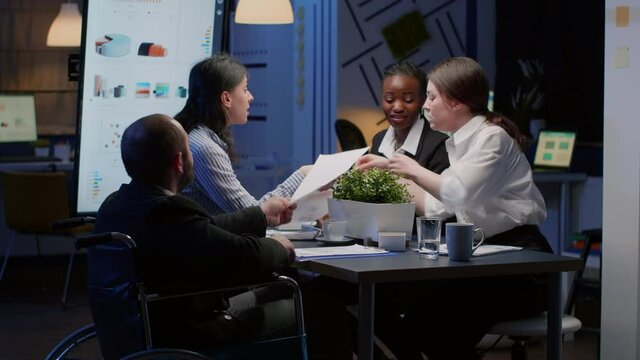 Handicapped paralyzed manager man in wheelchair sitting at conference table working in office meeting room. Businessman is ignored while coworkers analyzing company paperwork late at night