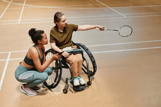 High angle view at young woman in wheelchair talking to coach or teammate during badminton practice at sports court, copy space