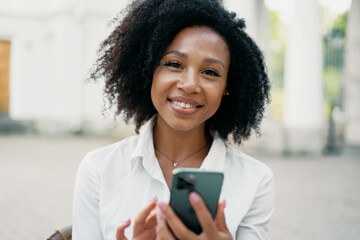 Portrait of a student typing a message on the phone. Blogger curly-haired young woman of African-American appearance smiles.