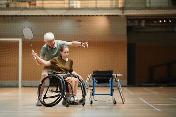 Full length portrait of senior coach training young woman in wheelchair during badminton practice...