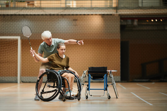 Full length portrait of senior coach training young woman in wheelchair during badminton practice in sports court, copy space