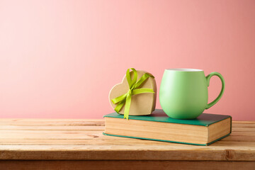 Green coffee cup with heart shape gift box and book  on wooden table over pink background