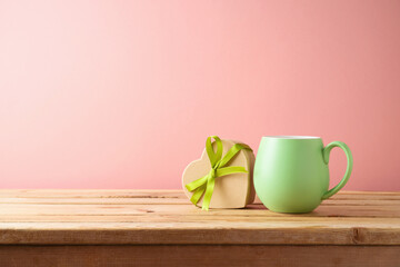Green coffee cup with heart shape gift box on wooden table over pink background