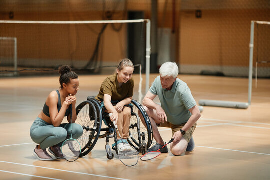 Full length portrait of mature coach talking to young woman in wheelchair during badminton practice in sports court, copy space