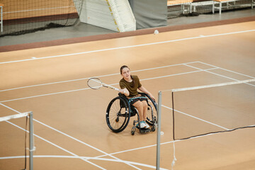 High angle view at young woman in wheelchair playing badminton during sports practice at indoor court, copy space - obrazy, fototapety, plakaty