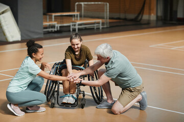 Full length portrait of young sportswoman in wheelchair huddling with team before match at indoor...