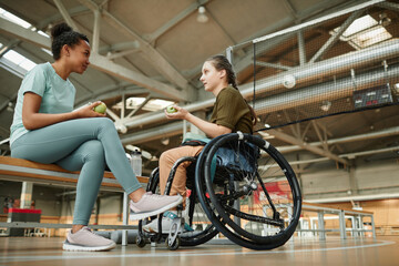 Obraz premium Full length portrait of young woman in wheelchair sharing lunch with friend at indoor sports court