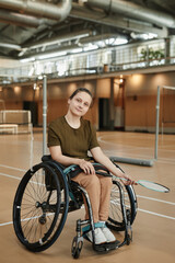 Fototapeta na wymiar Full length portrait of young woman in wheelchair looking at camera in indoor sports court