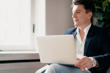 A professional manager writes a message to the client by email. An adult man in a suit with gray hair works in an office in a coworking.