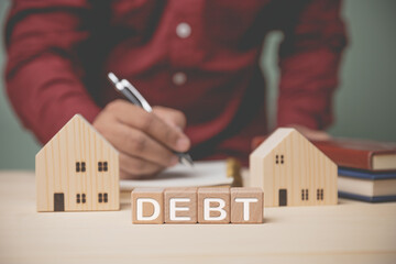 DEBT on wood blocks with houses model. Businessman holding a pen to record home expenses to...