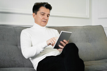 A business banker is a confident adult man, working on a laptop tablet in an apartment. Prints a message to its project partners. Sitting on a comfortable sofa, looking good.