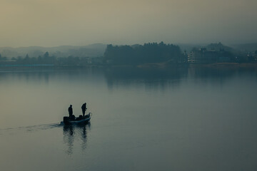 Fototapeta na wymiar Silhouette of two male fisherman sailing boat in the lake for fishing with landscape of wild forest in the background. Tree and man shadow reflect on water. Thick fog in morning with blue color tone.