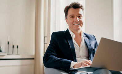 Fototapeta premium The professional manager smiles and writes a message to the client by email. An adult man in a suit with gray hair works in an office in a coworking.