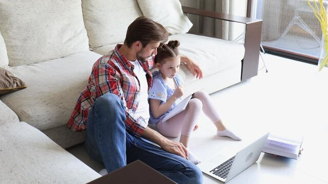 Smiling father and daughter sitting on floor in living room with laptop, teaching lessons.