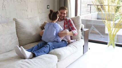 Cute little kid daughter laughing tickling playing with dad on sofa, happy father relaxing having fun with funny small child girl bonding enjoying leisure together in living room.