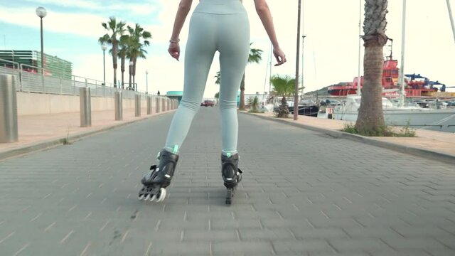 Slowmotion rear view showing sexy woman in leggings inline skating along pier promenade in summer.