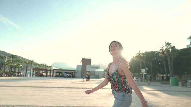 Slow motion track shot of happy smiling woman skating on promenade near the port during sunset in background
