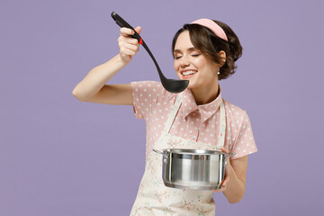 Young happy housewife housekeeper chef cook baker woman in pink apron holding soup stainless pan saucepan with ladle try taste isolated on pastel violet background studio Cooking food process concept - obrazy, fototapety, plakaty