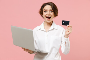 Surprised young employee business woman corporate lawyer in classic formal white shirt work in office hold using laptop pc computer credit bank card shopping online isolated on pastel pink background.