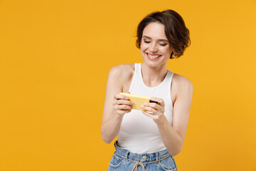 Young gambling woman 20s with bob haircut in white tank top shirt using play racing on mobile cell phone hold gadget smartphone for pc video games isolated on yellow color background studio portrait