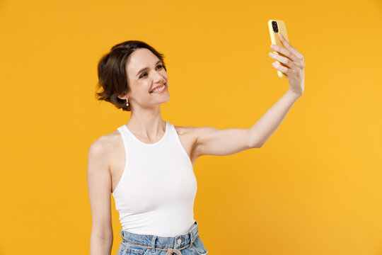 Young smiling happy friendly woman 20s with bob haircut wearing white tank top shirt doing selfie shot on mobile phone post photo on social network isolated on yellow color background studio portrait.