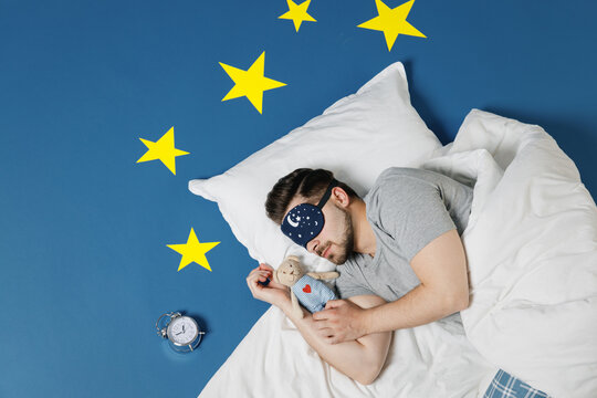 Top view young man in pajamas jam sleep mask resting relax at home lies wrap covered under blanket duvet hold teddy bear plush toy isolated on dark blue sky background Good mood night bedtime concept