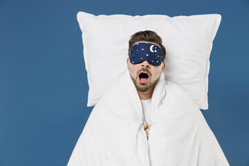 Young happy fun caucasian man 20s wearing pajamas jam sleep mask resting relaxing at home lies wrap...