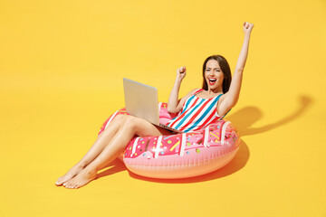 Full body length happy young sexy woman wear red blue swimsuit sit on inflatable ring work on pc laptop isolated on vivid yellow color background studio Summer hotel pool sea rest sun tan concept