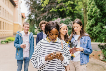Young african american woman in striped jacket with straight hair in striped with surprised or shocked emotion on face outdoors using smartphone with group of students behind on blurred background.