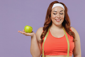 Young smiling vegeterian chubby overweight plus size big fat fit woman in red top measure tape warm up training look at green apple fruit isolated on purple background home gym Workout sport concept.