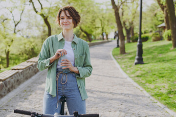 Young woman 20s in green jacket jeans riding bicycle bike in city spring park outdoors, drink clear...