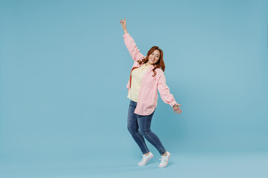 Full length young fun joyful smiling redhead chubby overweight woman 30s wearing in pink shirt jeans leaning back stand on toes with oustretched hands hands dance isolated on pastel blue background.