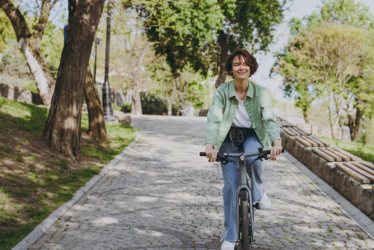 Young fun dreamful happy woman 20s wearing casual green jacket jeans riding bicycle bike on sidewalk in city spring park outdoors, look aside. People active urban healthy lifestyle cycling concept