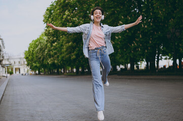 Full length young carefree woman in jeans clothes headphones listen to music walking stroll down green park alley outdoors run fast with outstretched raised up hands People urban lifestyle concept